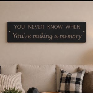 May include: A rectangular black wall sign featuring the engraved text "YOU NEVER KNOW WHEN You're making a memory" in a combination of uppercase and script fonts. The sign has a small circular hole on each side for mounting purposes.