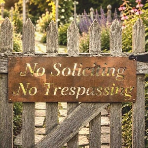 May include: A rectangular metal sign with a rusted finish mounted on a weathered wooden picket fence. The sign features laser-cut lettering that reads No Soliciting and No Trespassing in a clear, bold font, secured by screws at the corners.