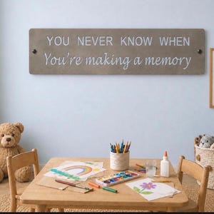 May include: A rectangular taupe wall sign featuring the white text "YOU NEVER KNOW WHEN You're making a memory" with two dark circular fasteners on the left and right sides. The sign is mounted horizontally on a light blue wall above a wooden table.