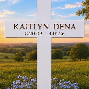 May include: A white cross monument with the name "KAITLYN DENA" and the dates "8.20.09 - 4.01.26" inscribed on it. The cross is set against a scenic backdrop of a field, trees, and a sunset sky, with blue flowers in the foreground.