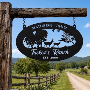May include: An oval black metal sign hanging from a wooden post by two chains. The sign features a cutout silhouette of trees, a deer, a doe, and birds. Text on the sign reads: MADISON, OHIO, Tucker's Ranch, and EST. 2000 in white lettering.