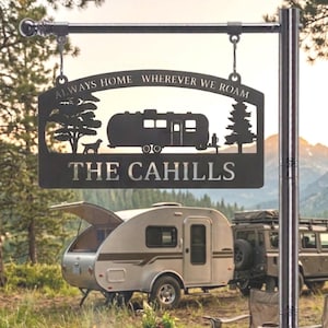 Puede incluir: Un letrero de metal negro colgado de un poste plateado. El letrero presenta una silueta de una caravana, un perro y &aacute;rboles, con el texto "ALWAYS HOME WHEREVER WE ROAM" y "THE CAHILLS" recortados de la placa de metal rectangular.