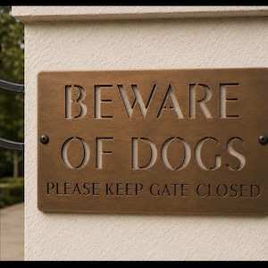May include: A rectangular bronze-colored metal sign with rounded corners, featuring the cut-out text BEWARE OF DOGS and the engraved text PLEASE KEEP GATE CLOSED. The sign is secured to a light-colored stone pillar with two visible black screws.