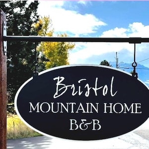以下が含まれることがあります： 「Bristol MOUNTAIN HOME B&B」と白い文字で書かれた黒い楕円形の看板。看板は黒い金属製のバーから吊り下げられています。背景には木々、山、雲のある青い空があります。