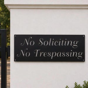 May include: A rectangular black metal sign mounted on a white pillar. The sign features cut-out lettering that reads No Soliciting and No Trespassing in a serif font. Four small silver screws secure the sign to the pillar at each corner.