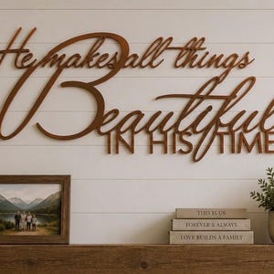 May include: A brown, laser-cut wooden wall sign featuring the cursive and block text "He makes all things Beautiful IN HIS TIME" mounted on a white horizontal plank wall above a wooden mantelpiece with decor items including plants and a lantern.