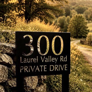 May include: Black metal address sign with the number "300", the text "Laurel Valley Rd PRIVATE DRIVE" cut out. The sign is mounted on a post next to a stone wall, with a natural, rural background.