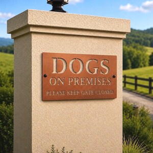 Pu&ograve; includere: Una targa rettangolare in metallo color ruggine montata su un pilastro beige testurizzato. La targa presenta lettere ritagliate che recitano DOGS ON PREMISES e un testo inciso sotto che dice PLEASE KEEP GATE CLOSED. Fissata con due viti scure.