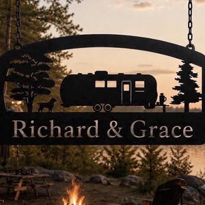 Puede incluir: Un letrero colgante de metal negro con la silueta de una caravana, un perro y dos &aacute;rboles. El texto "Richard & Grace" est&aacute; recortado en la parte inferior del letrero. El letrero est&aacute; suspendido por dos cadenas de metal en las esquinas.