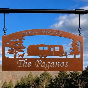 Puede incluir: Un letrero rectangular de metal color cobre que cuelga de una barra negra. El letrero presenta un dise&ntilde;o recortado de una caravana, un perro y dos &aacute;rboles. El texto RON & MAGGIE est&aacute; recortado arriba y The Paganos abajo.