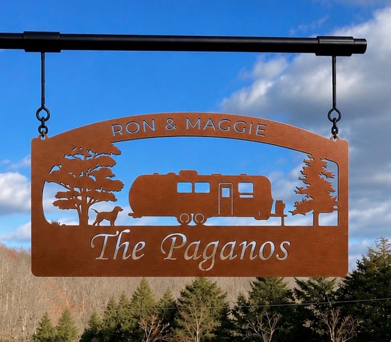 Puede incluir: Un letrero rectangular de metal color cobre que cuelga de una barra negra. El letrero presenta un dise&ntilde;o recortado de una caravana, un perro y dos &aacute;rboles. El texto RON & MAGGIE est&aacute; recortado arriba y The Paganos abajo.