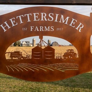 May include: A rust-colored metal sign featuring the text HETTERSIMER FARMS EST 2017. The sign has a cutout design depicting a farm scene with a combine harvester, grain silos, a tractor, and rows of crops, suspended by chains from a wooden post.