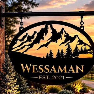 May include: An oval black metal sign hanging from a wooden post. The sign features a cutout silhouette of mountain peaks and pine trees. The text WESSAMAN is printed in gold lettering above the text EST. 2021, which is also in gold.