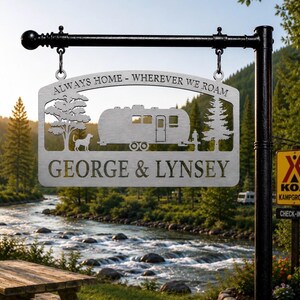 Puede incluir: Un letrero de metal plateado colgado de un poste negro, con un dise&ntilde;o recortado de una caravana, un &aacute;rbol, un pino y un perro. El texto dice "ALWAYS HOME - WHEREVER WE ROAM" y "GEORGE & LYNSEY" en una fuente negrita sin remates.