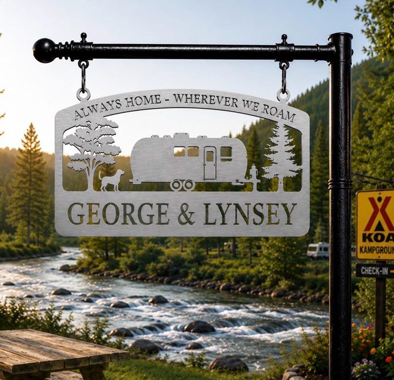 Puede incluir: Un letrero de metal plateado colgado de un poste negro, con un dise&ntilde;o recortado de una caravana, un &aacute;rbol, un pino y un perro. El texto dice "ALWAYS HOME - WHEREVER WE ROAM" y "GEORGE & LYNSEY" en una fuente negrita sin remates.
