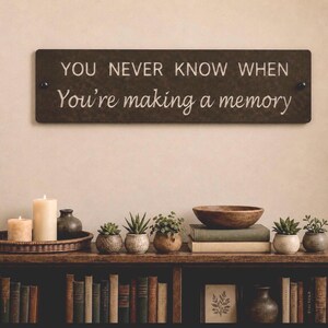 May include: A rectangular dark brown wall sign featuring the white text YOU NEVER KNOW WHEN You're making a memory. The sign is mounted with two black circular fasteners on each side and is positioned above a shelf filled with books and plants.
