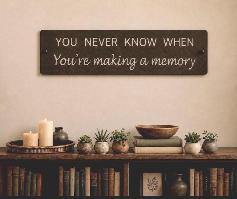 May include: A rectangular dark brown wall sign featuring the white text YOU NEVER KNOW WHEN You're making a memory. The sign is mounted with two black circular fasteners on each side and is positioned above a shelf filled with books and plants.