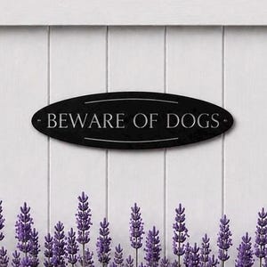 Può includere: Una targa ovale nera con scritte bianche che recita BEWARE OF DOGS. La targa presenta una sottile linea di bordo bianca sopra e sotto il testo e ha piccoli fori di montaggio circolari sulle estremità sinistra e destra della forma ovale.
