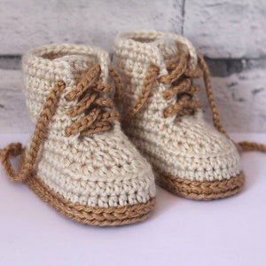 May include: A pair of crocheted baby booties with brown laces and a brown trim. The booties are made with a white yarn.