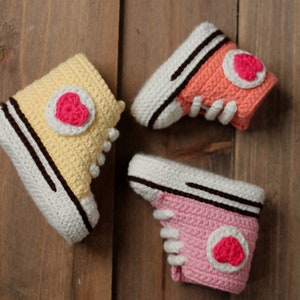 Peut inclure: Trois paires de chaussons en crochet pour bébé en rose, jaune et orange. Chaque chausson a un cœur blanc avec un centre rose sur le côté.