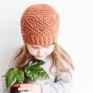 May include: A young person wearing a brown crocheted beanie hat and a grey sweater holds a potted plant with green leaves.