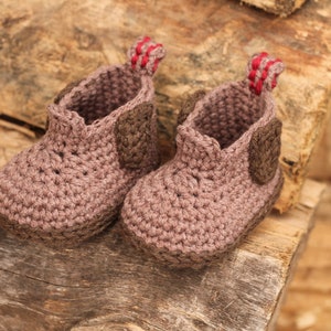 Puede incluir: Dos botines de beb&eacute; de ganchillo marr&oacute;n con ribetes rojos. Los botines est&aacute;n hechos con un punto texturizado y tienen un dise&ntilde;o sencillo.