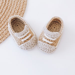 Puede incluir: Un par de zapatos de bebé de ganchillo beige y blanco con un diseño sencillo. Los zapatos tienen una punta redonda y una suela plana.