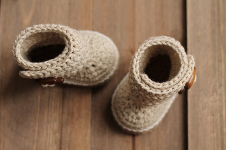K&ouml;nnte beinhalten: Zwei geh&auml;kelte Babyschuhe in beige. Die Schuhe haben ein schlichtes Design und sind aus weichem Garn gefertigt.
