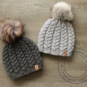 Puede incluir: Dos gorros de ganchillo con pompones de piel sintética. Los gorros son grises y gris oscuro con un patrón trenzado. Los gorros tienen una etiqueta de cuero marrón con el texto "Inventorium Crochet Pattern".