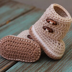 K&ouml;nnte beinhalten: Ein Paar geh&auml;kelte Babyschuhe mit braunen Kn&ouml;pfen. Die Schuhe sind beige und braun mit einem wei&szlig;en Rand.
