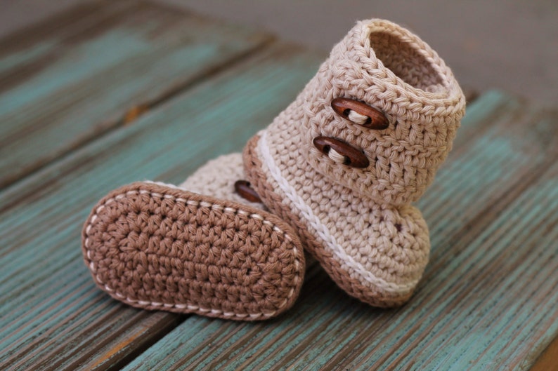K&ouml;nnte beinhalten: Ein Paar geh&auml;kelte Babyschuhe mit braunen Kn&ouml;pfen. Die Schuhe sind beige und braun mit einem wei&szlig;en Rand.