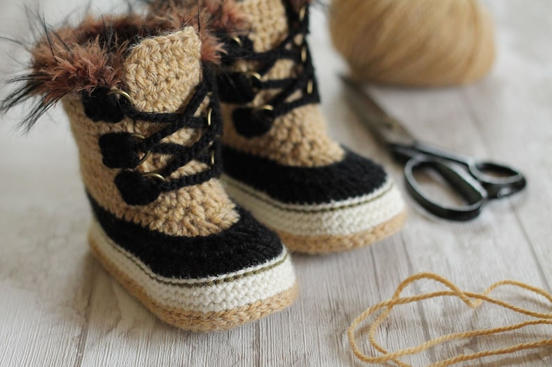 May include: A pair of crocheted baby booties with a black and white trim, brown fur trim, and brown laces. The booties are sitting on a white wooden surface with a ball of yarn and scissors.