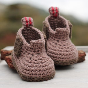Puede incluir: Un par de botitas de beb&eacute; de crochet marr&oacute;n con detalles rojos. Las botitas tienen un dise&ntilde;o sencillo y est&aacute;n hechas con un hilo suave y texturizado.