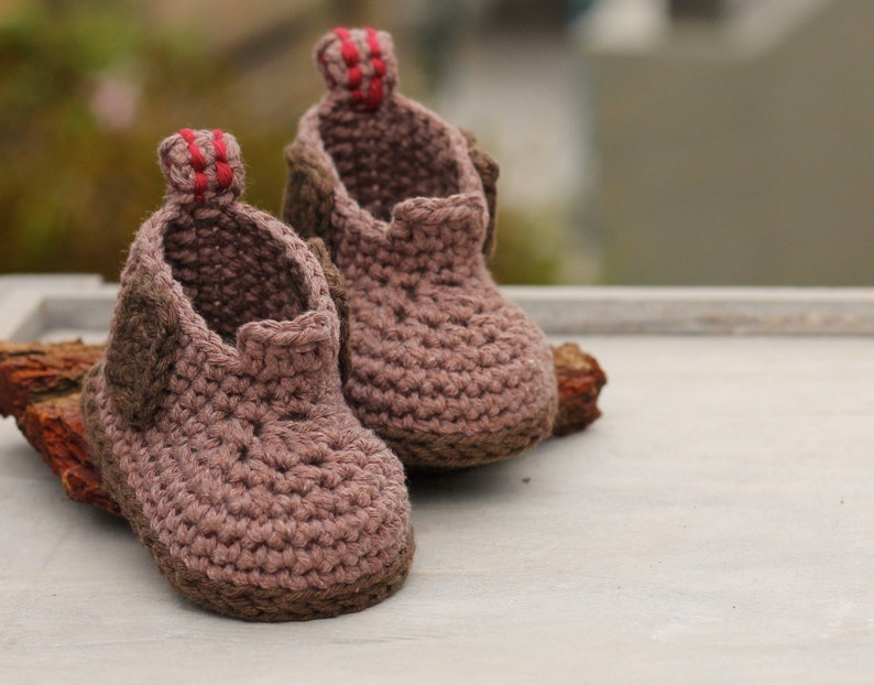 Puede incluir: Un par de botitas de beb&eacute; de crochet marr&oacute;n con ribetes rojos. Las botitas est&aacute;n hechas con un hilo suave y texturizado y tienen un dise&ntilde;o simple y cl&aacute;sico.