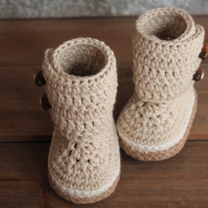 K&ouml;nnte beinhalten: Ein Paar beige gestrickte Babyschuhe mit braunen Kn&ouml;pfen. Die Schuhe haben einen wei&szlig;en Saum am oberen Rand.