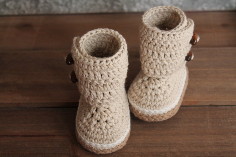 K&ouml;nnte beinhalten: Ein Paar beige gestrickte Babyschuhe mit braunen Kn&ouml;pfen. Die Schuhe haben einen wei&szlig;en Saum am oberen Rand.