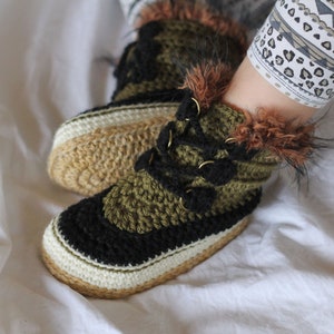 May include: Close-up of a pair of crocheted slippers with black, green, and white yarn. The slippers have a faux fur trim and a gold buckle detail.