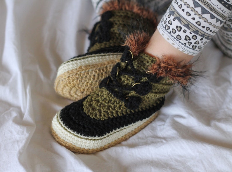 May include: Close-up of a pair of crocheted slippers with black, green, and white yarn. The slippers have a faux fur trim and a gold buckle detail.