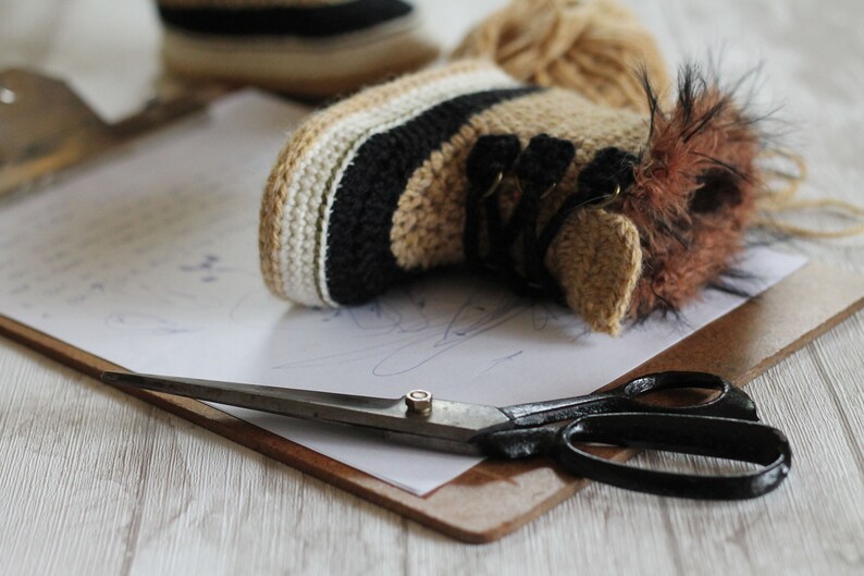 May include: A pair of crocheted baby booties with brown fur trim, a pair of scissors, and a piece of paper on a clipboard.