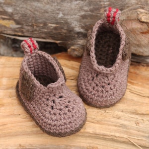 Puede incluir: Un par de botitas de beb&eacute; de crochet marr&oacute;n con ribetes rojos. Las botitas tienen un dise&ntilde;o sencillo y est&aacute;n hechas con un hilo suave.