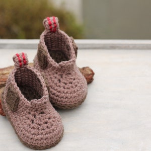 Puede incluir: Un par de botitas de beb&eacute; de crochet marr&oacute;n con ribetes rojos. Las botitas tienen una aplicaci&oacute;n de fieltro marr&oacute;n en la punta.