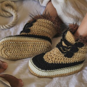 May include: Crocheted tan and black slippers with faux fur trim. The slippers have a lace-up design with black laces and gold-colored eyelets.