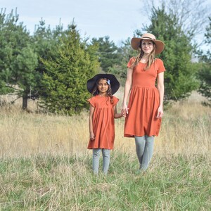 May include: Two people wearing matching rust-colored dresses and hats stand in a field of tall grass. The person in the front is wearing a black hat and the person in the back is wearing a tan hat.
