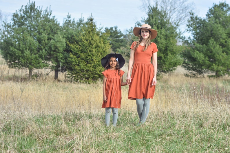 May include: Two people wearing matching rust-colored dresses and hats stand in a field of tall grass. The person in the front is wearing a black hat and the person in the back is wearing a tan hat.