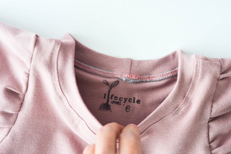 May include: A pink t-shirt with a small, black, printed label that says "lifecycle apparel 6" and a small, black, printed image of a plant.