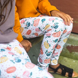May include: Two pairs of leggings with a white background and a pattern of orange, pink, and yellow pumpkins, green leaves, and white polka dots.