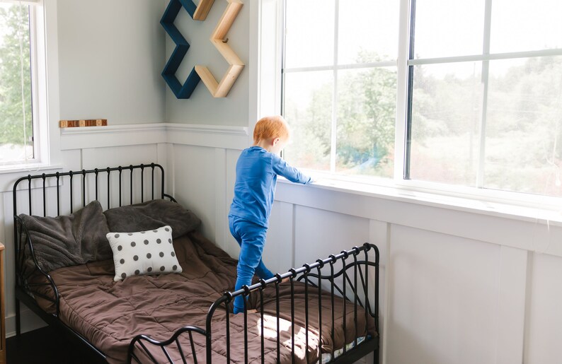 Little boy in organic cotton pajamas stands on bed near window