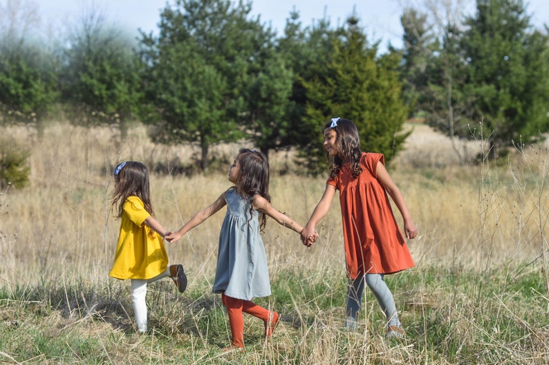May include: Three girls in dresses hold hands and walk through a field of tall grass. The girl in the front is wearing a yellow dress, the middle girl is wearing a blue dress, and the girl in the back is wearing a red dress.