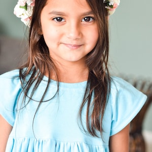 May include: A young girl with long brown hair wears a light blue dress and a flower crown with pink and white flowers.