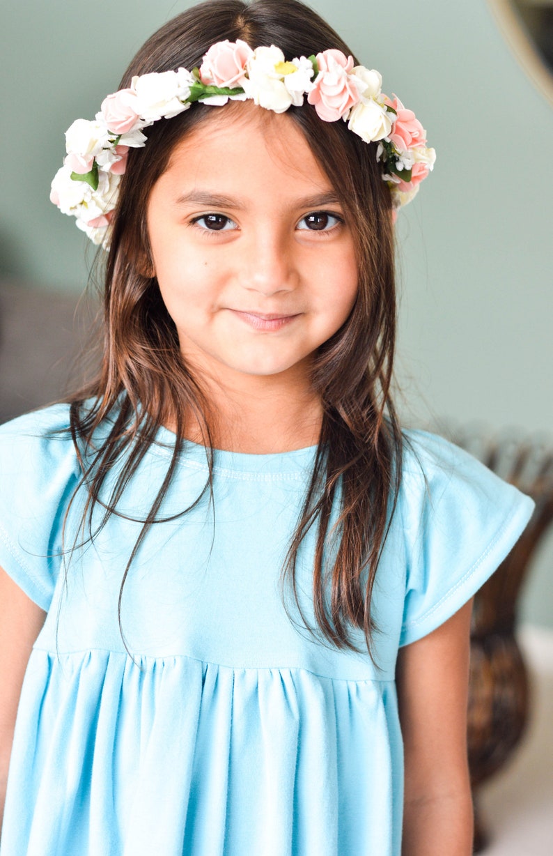 May include: A young girl with long brown hair wears a light blue dress and a flower crown with pink and white flowers.